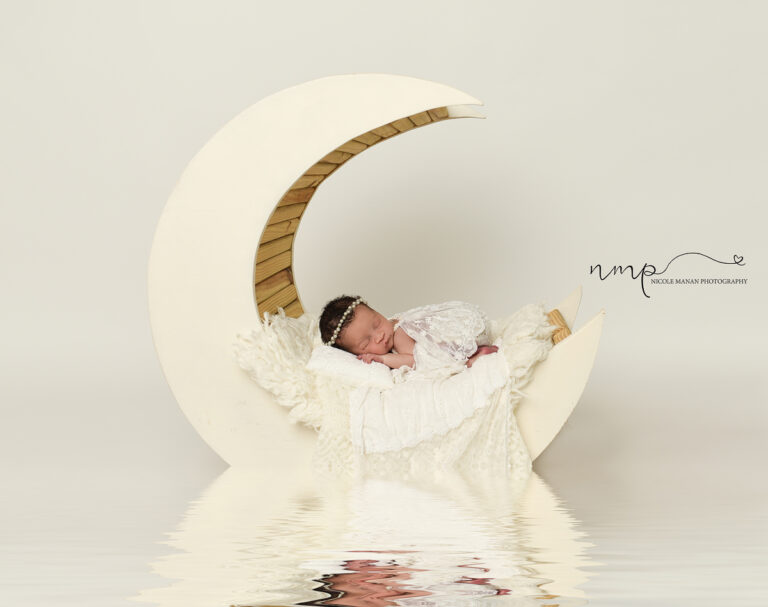 A sleeping newborn baby girl in a white lace outfit on a white moon prop during her Columbus GA Newborn session.