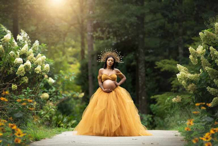 Pregnant woman wearing a goddess crown and beautiful mustard maternity dress in a lush garden setting at golden hour during her Columbus GA maternity session
