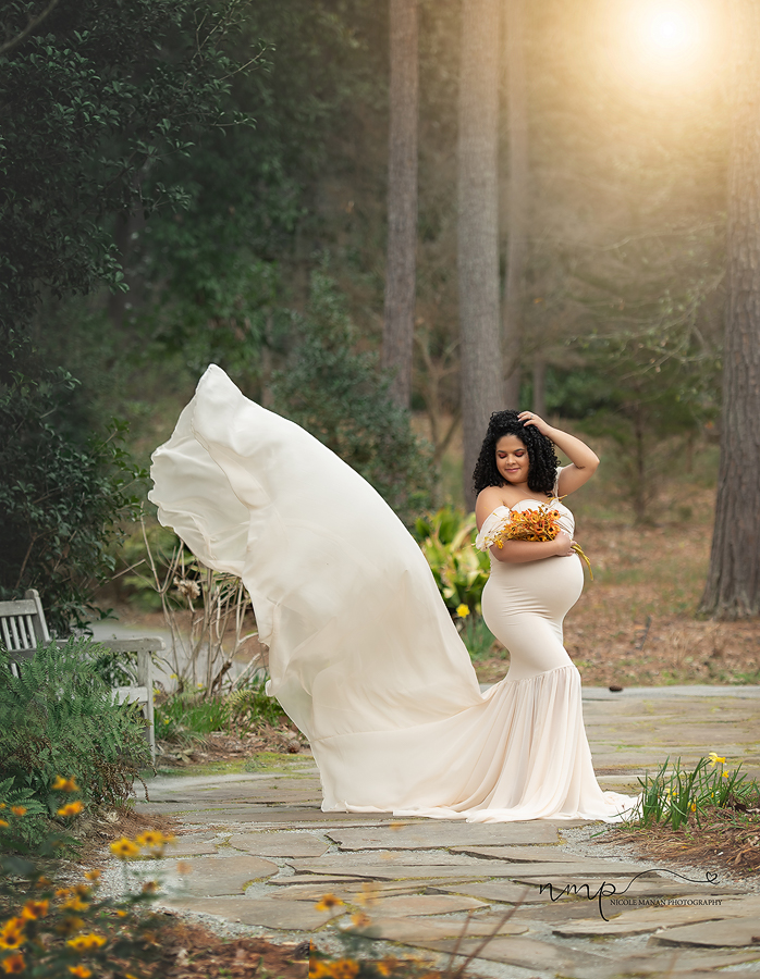 Pregnant woman in a white flowy dress with a tossing train holding flowers at her Callaway Garden outdoor maternity session.