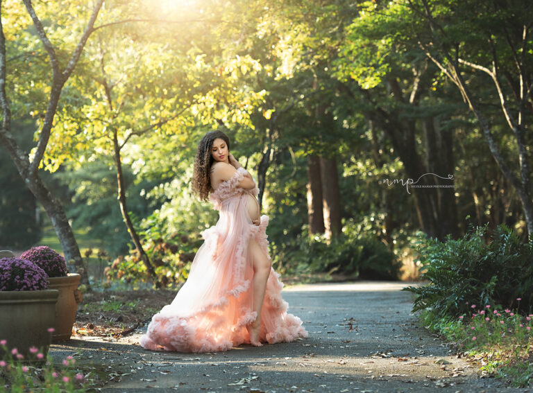 A pregnant woman during her maternity session in a beautiful flowy pink dress standing in golden light in a lushes park.