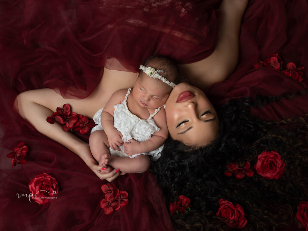 A mother and her newborn baby girl covered in red fabric and surrounded by red flowers, they are lying down in a loving pose during their Columbus GA newborn session.