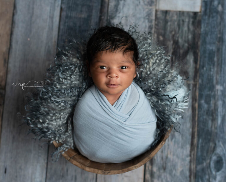 A newborn baby boy swaddles in blue in a wooden bowl with his eyes wide open and a little smile durin his Columbus GA newborn session.