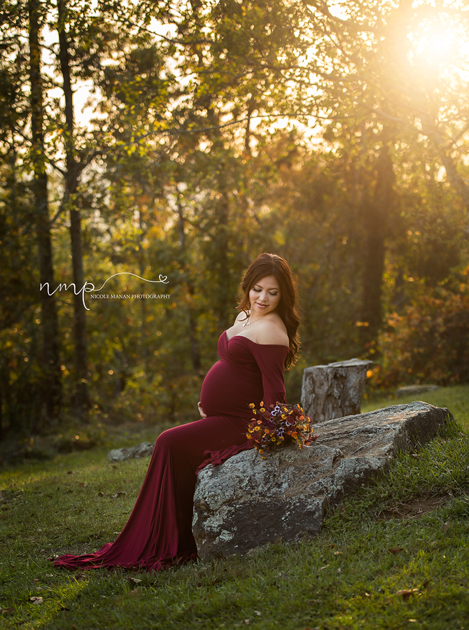 A pregnant woman in a burgundy dress at golden hour sitting on a rock during her Pine Mountain maternity session.