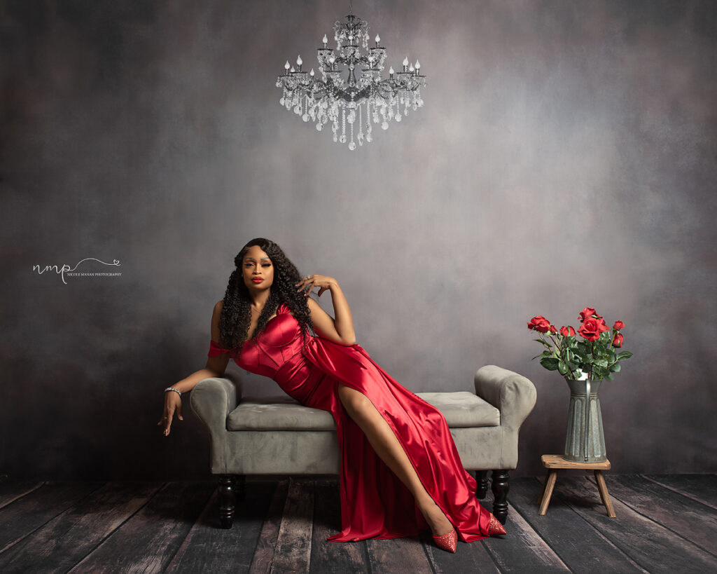 A woman in a beautiful red dress sitting on a sofa with roses next to her and a beautiful chandelier