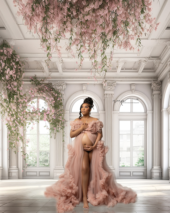 A pregnant black woman in a pink ruffle dress in a large white room decorated with pink flowers during her Columbus GA maternity session.