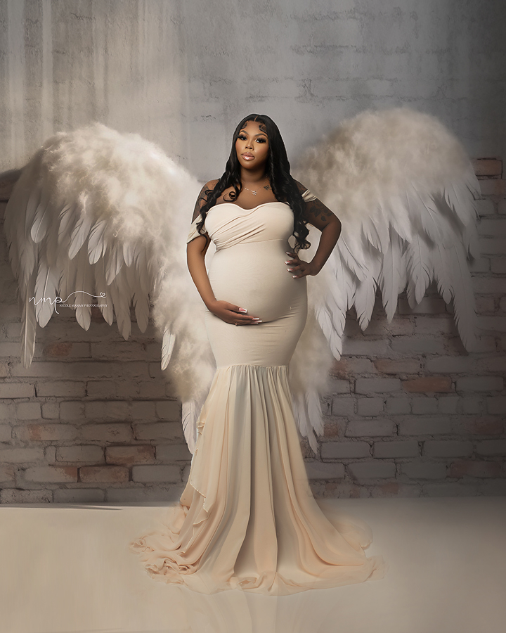 Pregnant black woman in a white dress and white giant angel wings in a studio maternity session.