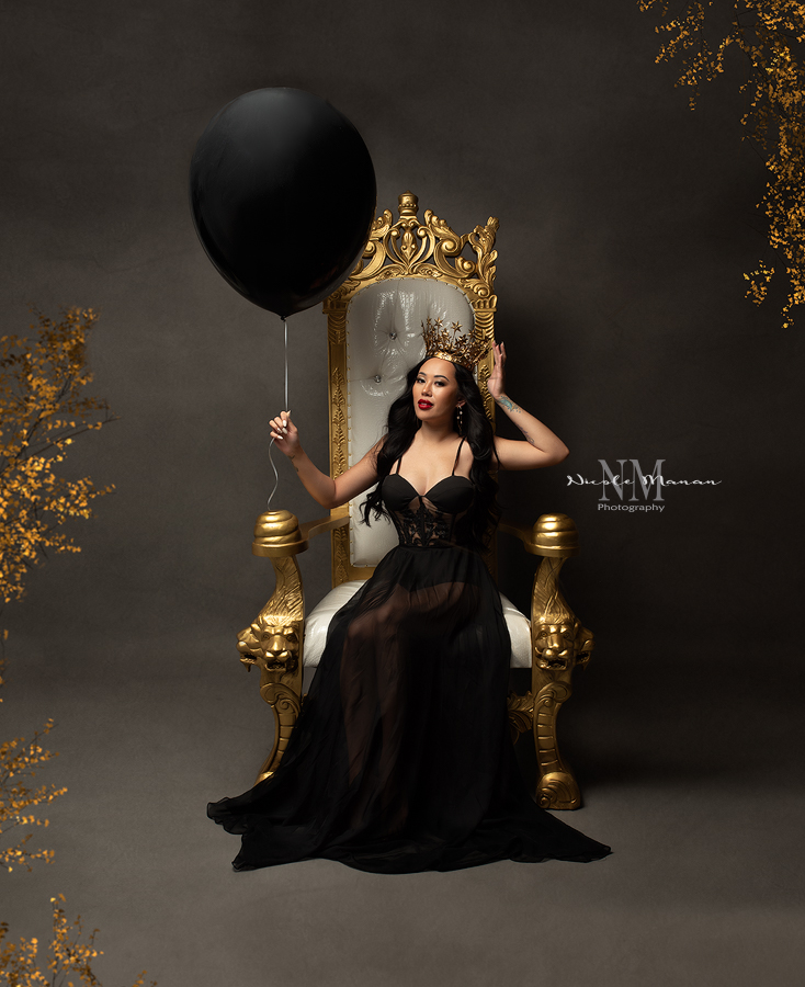 A woman in a black dress holding a black balloon and sitting on a golden throne with a golden crown during her Columbus Ga boudoir birthday session.