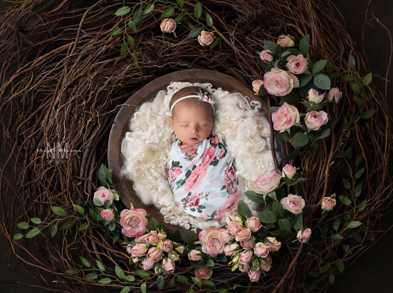 A newborn baby girl swaddled in a floral swaddle in a bowl surrounded by pink roses